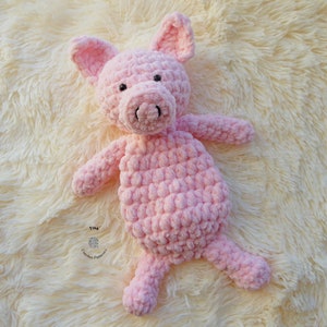 May include: A pink crocheted pig stuffed animal lying on a white fluffy surface. The pig has a small black nose and black eyes.
