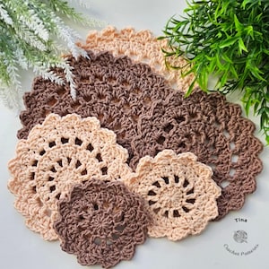 May include: A collection of handmade crochet doilies in various sizes and shades of brown and beige. The doilies have a floral design with scalloped edges. The image includes green and white foliage in the background.