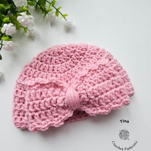 May include: A pink crocheted turban-style hat with a bow detail. The hat is handmade and has a textured appearance. The image also includes the text "Tina" and "Crochet Patterns".