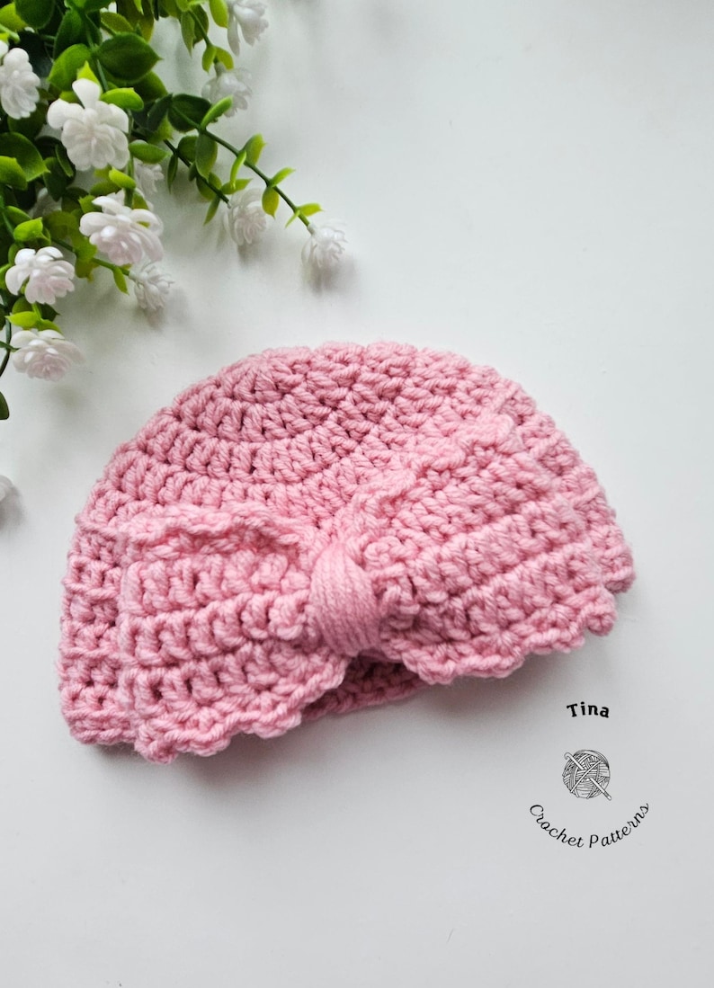 May include: A pink crocheted turban-style hat with a bow detail. The hat is handmade and has a textured appearance. The image also includes the text "Tina" and "Crochet Patterns".