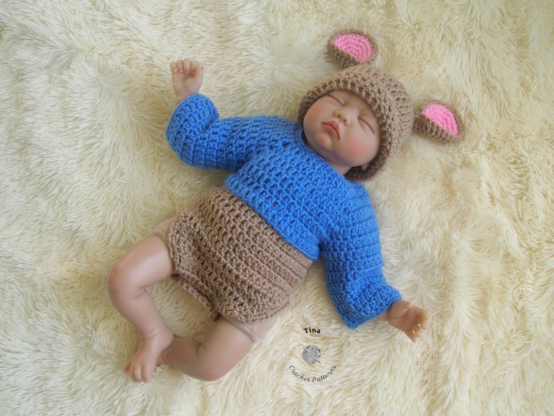 May include: A crocheted baby outfit in blue and brown with a matching crocheted hat featuring pink ears. The hat has a brown crocheted brim and pink crocheted ears. The outfit has a blue crocheted top and a brown crocheted bottom. The outfit is laid out on a white fluffy surface.
