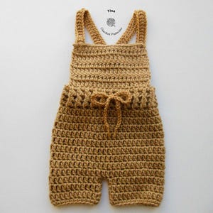 Op de afbeelding: Een gehaakte baby romper in een lichtbruine kleur met een strik in de taille. De romper heeft bandjes die in de rug kruisen.