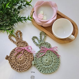 Può includere: Una composizione a tema pasquale con due sottobicchieri a forma di coniglietto all'uncinetto, uno marrone e uno verde, ciascuno con un fiocco rosa. Un portacandele rosa a forma di rosa con una candela bianca e una tazza da tè su un vassoio di legno.