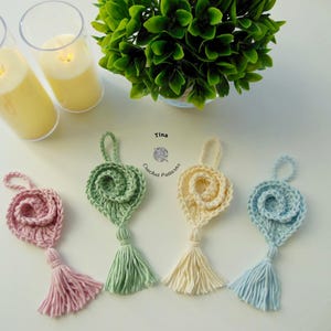 May include: Four crocheted flower ornaments with tassels in pink, green, cream, and blue. The ornaments are hanging from a white surface. Tina Crochet Patterns is written on the surface.