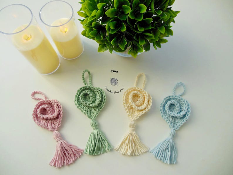 May include: Four crocheted flower ornaments with tassels in pink, green, cream, and blue. The ornaments are hanging from a white surface. Tina Crochet Patterns is written on the surface.