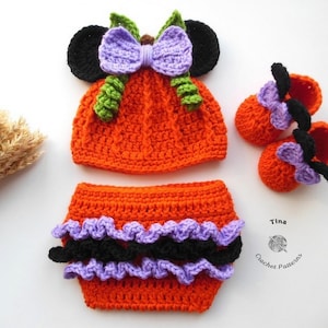 Può includere: Un set per neonati all'uncinetto fatto a mano in arancione, nero e viola. Il set comprende un cappello con orecchie nere e un fiocco viola, una mutandina con volant e scarpine abbinate. Il set è progettato per assomigliare a una zucca.