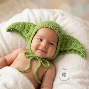 Può includere: Un bambino sorridente indossa un cappello verde all'uncinetto con grandi orecchie a punta e una chiusura con fiocco. Il bambino è sdraiato su una superficie bianca e soffice, avvolto in una coperta bianca. Il cappello è verde chiaro.
