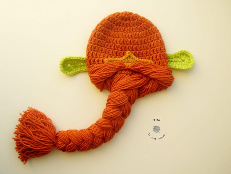 May include: A crocheted hat with a long braided tail, featuring a green ear on each side and a yellow star on the top. The hat is orange and the tail is braided in a brown color.