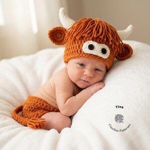 May include: A newborn baby wearing a crocheted orange Highland cow costume, including a hat with horns and a matching diaper cover with a tail. The baby is lying on a white surface.