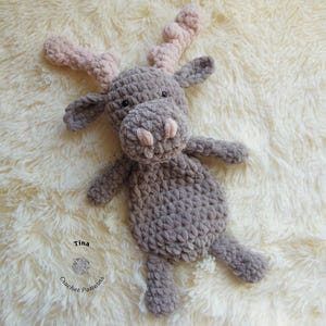 Puede incluir: Un alce de crochet gris con cuernos rosas. El alce está acostado sobre una superficie blanca y esponjosa. El texto "Tina Crochet Patterns" es visible en la esquina inferior izquierda de la imagen.