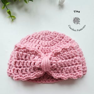 May include: A handmade, pink crochet turban-style hat. The hat features a knotted detail at the front and is made with a textured stitch. The image includes the text "Tina Crochet Patterns".