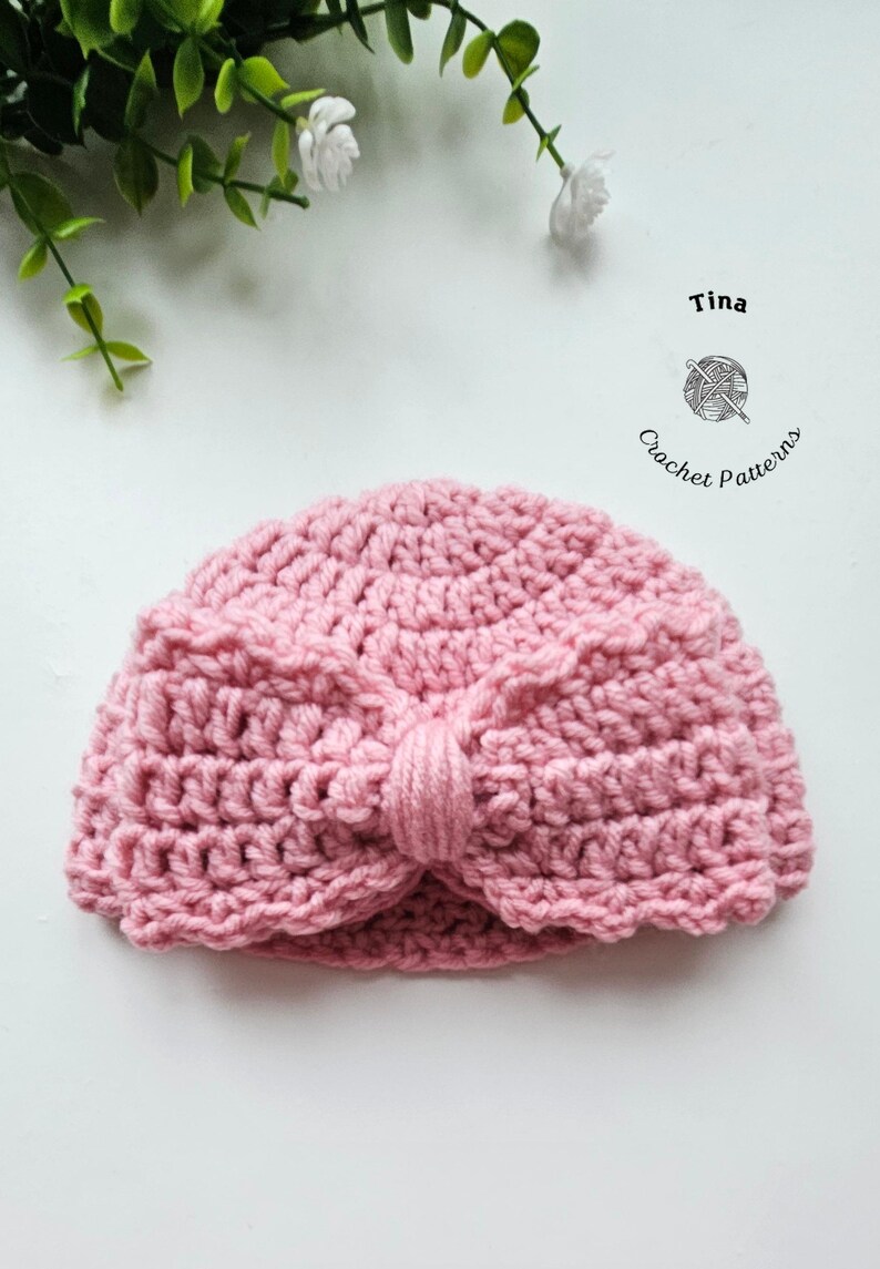 May include: A handmade, pink crochet turban-style hat. The hat features a knotted detail at the front and is made with a textured stitch. The image includes the text "Tina Crochet Patterns".