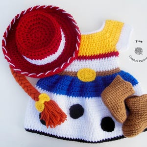 May include: Crocheted baby outfit resembling a cowgirl costume, featuring a red hat with white stitching, a dress with yellow, white, and blue sections, and brown boots. The outfit includes a braided hair piece and a yellow belt buckle.