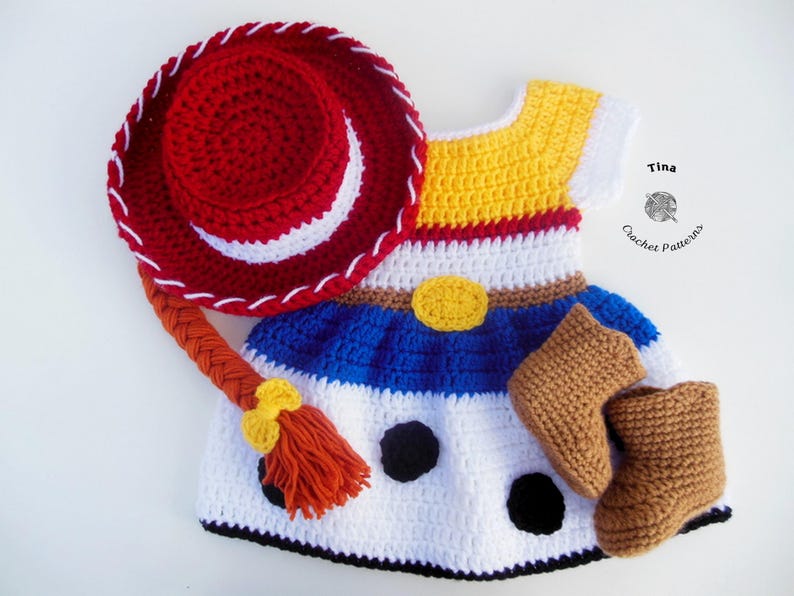May include: Crocheted baby outfit resembling a cowgirl costume, featuring a red hat with white stitching, a dress with yellow, white, and blue sections, and brown boots. The outfit includes a braided hair piece and a yellow belt buckle.