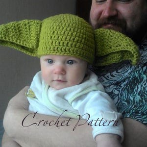 May include: A green crocheted hat with large ears, resembling a popular fictional character, is worn by a baby being held by an adult.