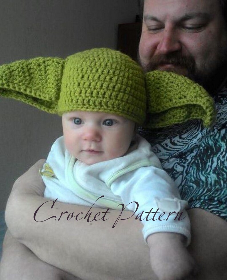 May include: A green crocheted hat with large ears, resembling a popular fictional character, is worn by a baby being held by an adult.
