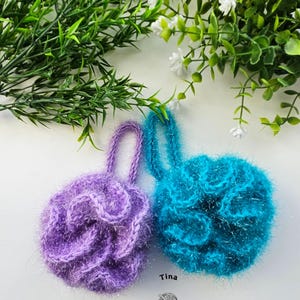 May include: Two handmade crochet bath scrubbies, one purple and one teal, with loop handles. The scrubbies have a textured, fluffy appearance. The image includes green foliage and the text "Tina Crochet Patterns".