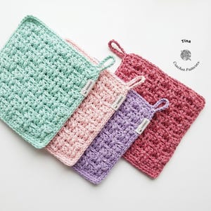 Puede incluir: Cuatro paños de crochet hechos a mano en tonos verde menta, rosa, morado y burdeos. Cada paño cuadrado presenta un patrón de tejido texturizado y un lazo para colgar. Los paños tienen una pequeña etiqueta que dice "handmade".