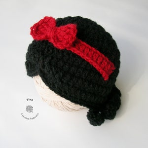 May include: A black crocheted hat with a red bow on top. The hat is shaped like a head with a ponytail at the back. The hat is sitting on a ball of yarn.