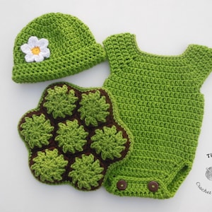 May include: A green crochet set consisting of a beanie with a white flower, a sleeveless romper with two brown buttons, and a textured brown and green turtle shell-shaped piece. The text Tina Crochet Patterns is visible in the bottom right corner.