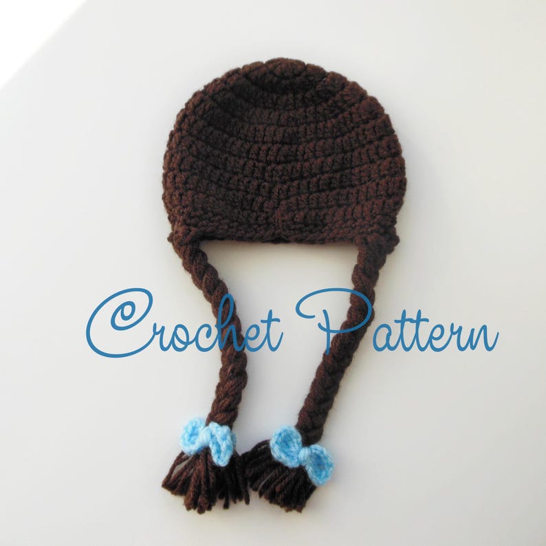 Może przedstawiać: Brązowy kapelusz na szydełku z dwoma długimi brązowymi warkoczami i niebieskimi kokardkami na końcach. "Crochet Pattern" jest napisane niebieskim kolorem pod kapeluszem.