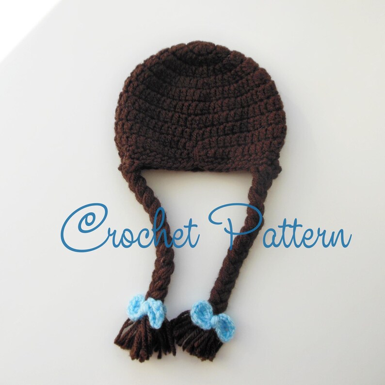 Może przedstawiać: Brązowy kapelusz na szydełku z dwoma długimi brązowymi warkoczami i niebieskimi kokardkami na końcach. "Crochet Pattern" jest napisane niebieskim kolorem pod kapeluszem.