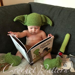 May include: A baby wearing a green crocheted Yoda hat and matching booties is reading a book titled "Star Wars ABC-3PO".