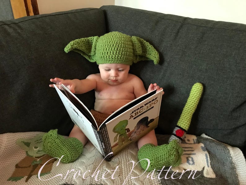 May include: A baby wearing a green crocheted Yoda hat and matching booties is reading a book titled "Star Wars ABC-3PO".