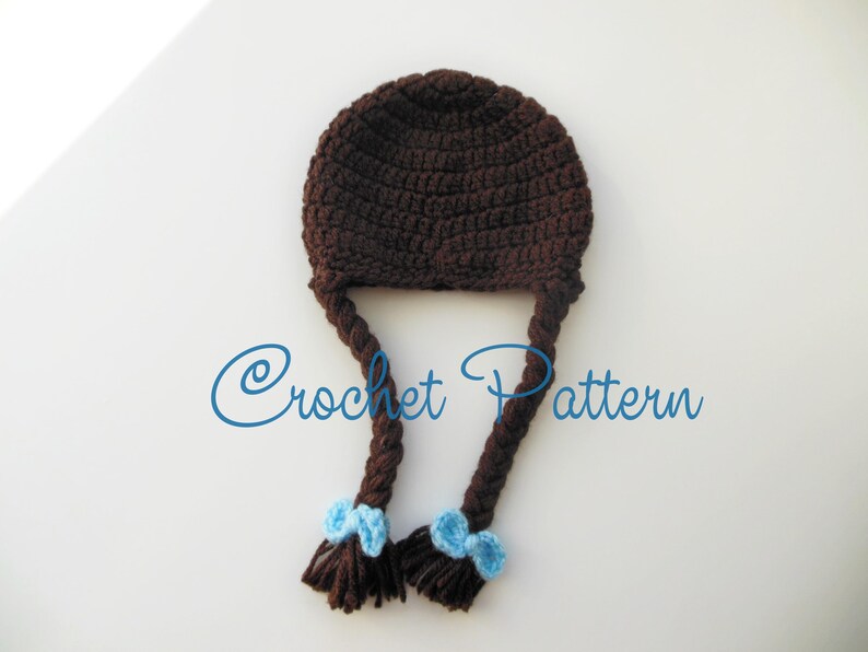 Może przedstawiać: Brązowa czapka na szydełku z dwoma długimi warkoczami i niebieskimi kokardkami na końcach. "Crochet Pattern" jest napisane niebieskim kolorem na białym tle.