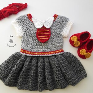 May include: A gray crocheted baby dress with red and gold accents. The dress has a white crocheted collar and a red crocheted bow. The dress is paired with red crocheted shoes with gold bows.