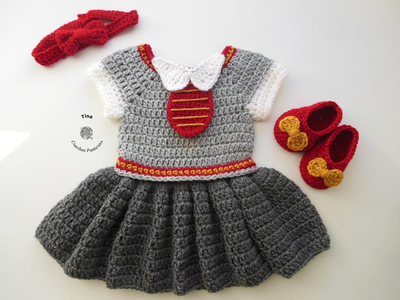 May include: A gray crocheted baby dress with red and gold accents. The dress has a white crocheted collar and a red crocheted bow. The dress is paired with red crocheted shoes with gold bows.