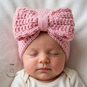 Op de afbeelding: Een baby draagt een lichtroze gehaakte strikmuts. De handgemaakte muts heeft een getextureerde steek en een grote strik. De baby draagt een witte romper. De tekst "Tina Crochet Patterns" staat linksonder.