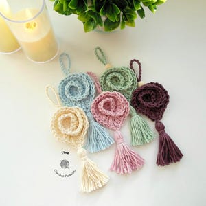 May include: Five crocheted flower ornaments with tassels in various colors, including light blue, cream, pink, green, and burgundy. The ornaments are arranged on a white surface. The text "Tina Crochet Patterns" is visible in the lower left corner of the image.