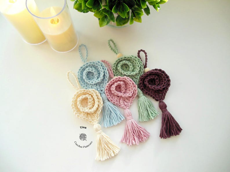 May include: Five crocheted flower ornaments with tassels in various colors, including light blue, cream, pink, green, and burgundy. The ornaments are arranged on a white surface. The text "Tina Crochet Patterns" is visible in the lower left corner of the image.