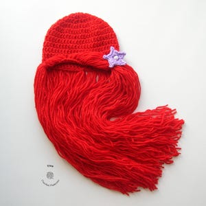 May include: Red crocheted hat with a long red yarn tail, resembling hair, and a purple crocheted starfish on top.
