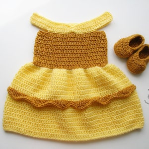 K&ouml;nnte beinhalten: Ein gelbes und braunes geh&auml;keltes Babykleid mit einem R&uuml;schenrock und passenden geh&auml;kelten Schuhen. Das Kleid hat einen einfachen, schulterfreien Ausschnitt.