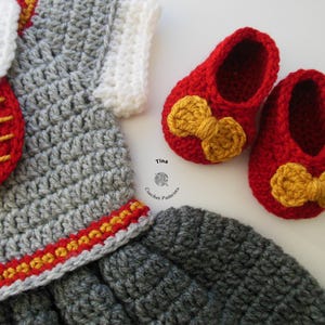 May include: A gray crocheted baby dress with a red and yellow trim, a red crocheted bow, and a red crocheted purse with a yellow stripe. The dress is paired with red crocheted baby shoes with yellow bows.