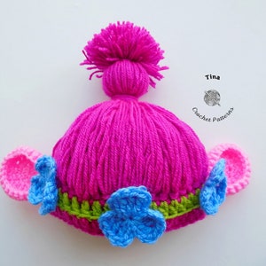 May include: Pink crocheted hat with blue and green flower accents and pink ear flaps. The hat is styled to look like a Troll doll with a pink pom-pom on top. Tina Crochet Patterns.