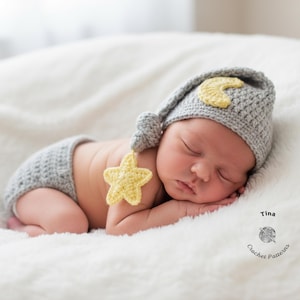 Op de afbeelding: Een pasgeboren baby met een gehaakte grijze muts met een gele maan en bijpassende ster. De baby draagt ook gehaakte grijze shorts en ligt op een zacht, wit oppervlak.