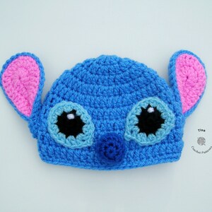 May include: Blue crocheted hat with pink ears and black eyes, resembling a cartoon character. The hat has a blue nose and white accents on the eyes.