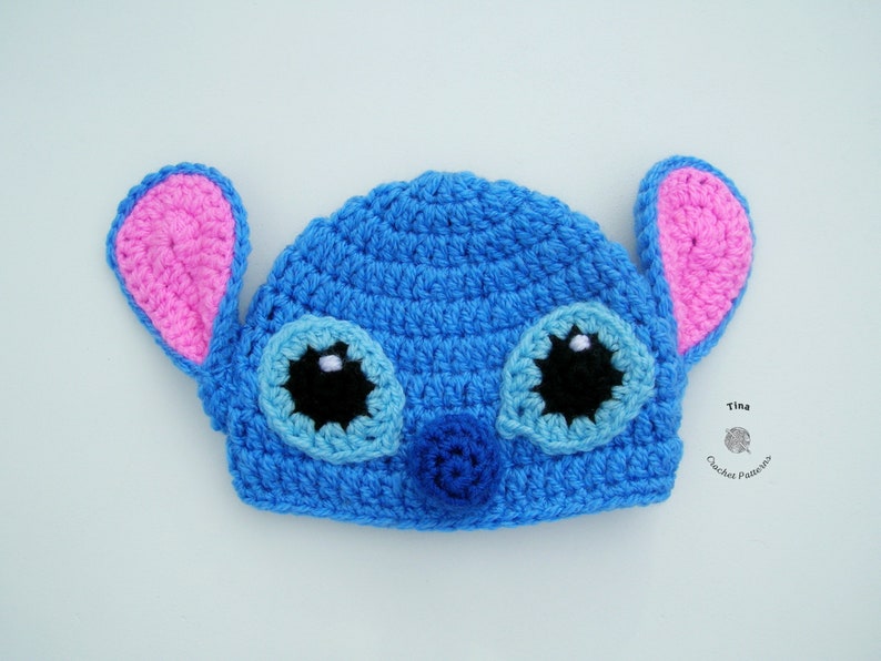 May include: Blue crocheted hat with pink ears and black eyes, resembling a cartoon character. The hat has a blue nose and white accents on the eyes.