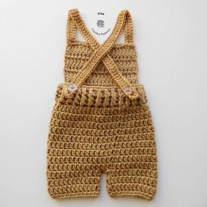 Op de afbeelding: Een gehaakte baby romper in een lichtbruine kleur met een kruisband ontwerp en twee knopen. De romper is gemaakt met een textuur steekpatroon.
