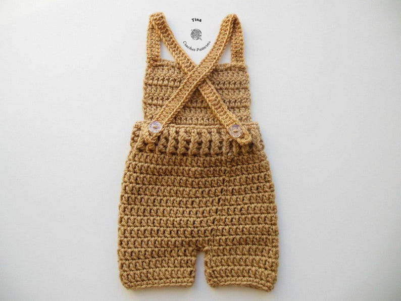 Op de afbeelding: Een gehaakte baby romper in een lichtbruine kleur met een kruisband ontwerp en twee knopen. De romper is gemaakt met een textuur steekpatroon.