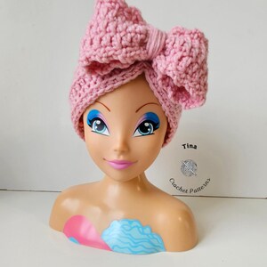 May include: A light pink, crocheted headband with a large bow detail. The headband is displayed on a mannequin head with blue eye makeup and pink lips. The text "Tina Crochet Patterns" is visible.