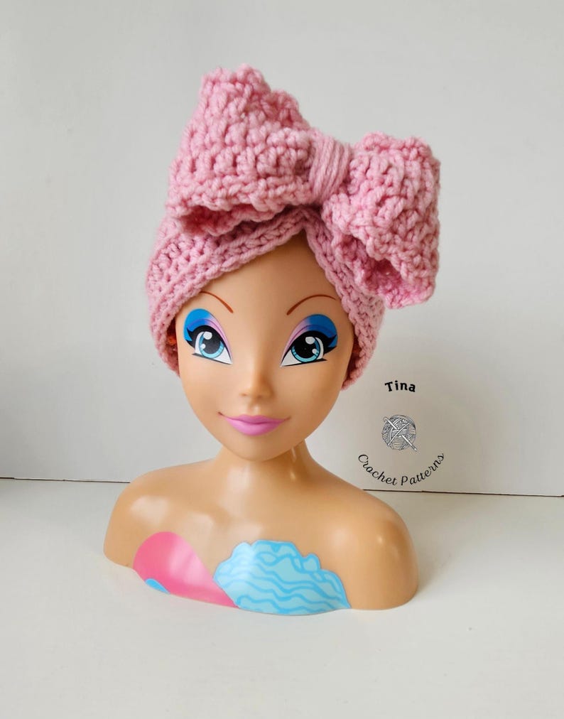 May include: A light pink, crocheted headband with a large bow detail. The headband is displayed on a mannequin head with blue eye makeup and pink lips. The text "Tina Crochet Patterns" is visible.