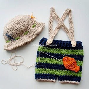 May include: A crocheted baby outfit with a beige hat, blue and green striped overalls, and a small orange fish appliqué. The outfit is perfect for a newborn photo shoot.