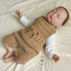 Op de afbeelding: Een bruine gehaakte baby romper met een strikdetail. De romper is gemaakt met een eenvoudig steekpatroon en heeft een ontspannen pasvorm. De romper wordt gedragen door een babypop.
