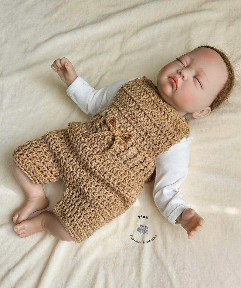 Op de afbeelding: Een bruine gehaakte baby romper met een strikdetail. De romper is gemaakt met een eenvoudig steekpatroon en heeft een ontspannen pasvorm. De romper wordt gedragen door een babypop.
