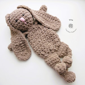 May include: A brown crocheted bunny plush toy with long floppy ears. The bunny is lying on its back with its legs and arms outstretched.