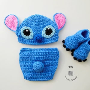 May include: A blue crocheted baby hat, diaper cover, and booties set featuring a Stitch character design. The hat has pink ears, black eyes, and a blue nose. The booties have black toes.